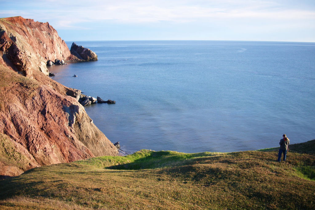 A man standing on the edge of a red, sandstone sea cliff, looking at the sea, on a sunny day.