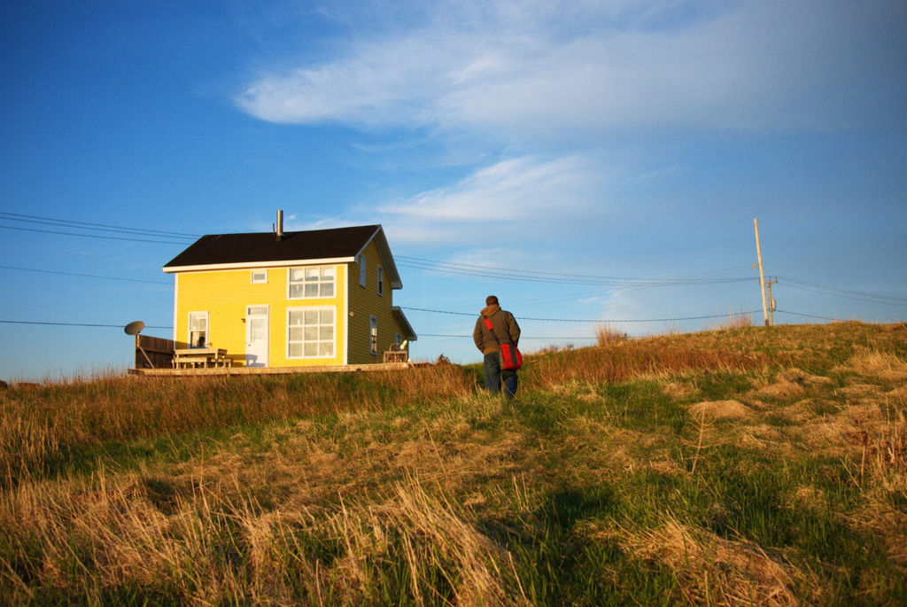 A man walking away from the camerea towards a yellow saltbox house in a wide, open space on a sunny day.