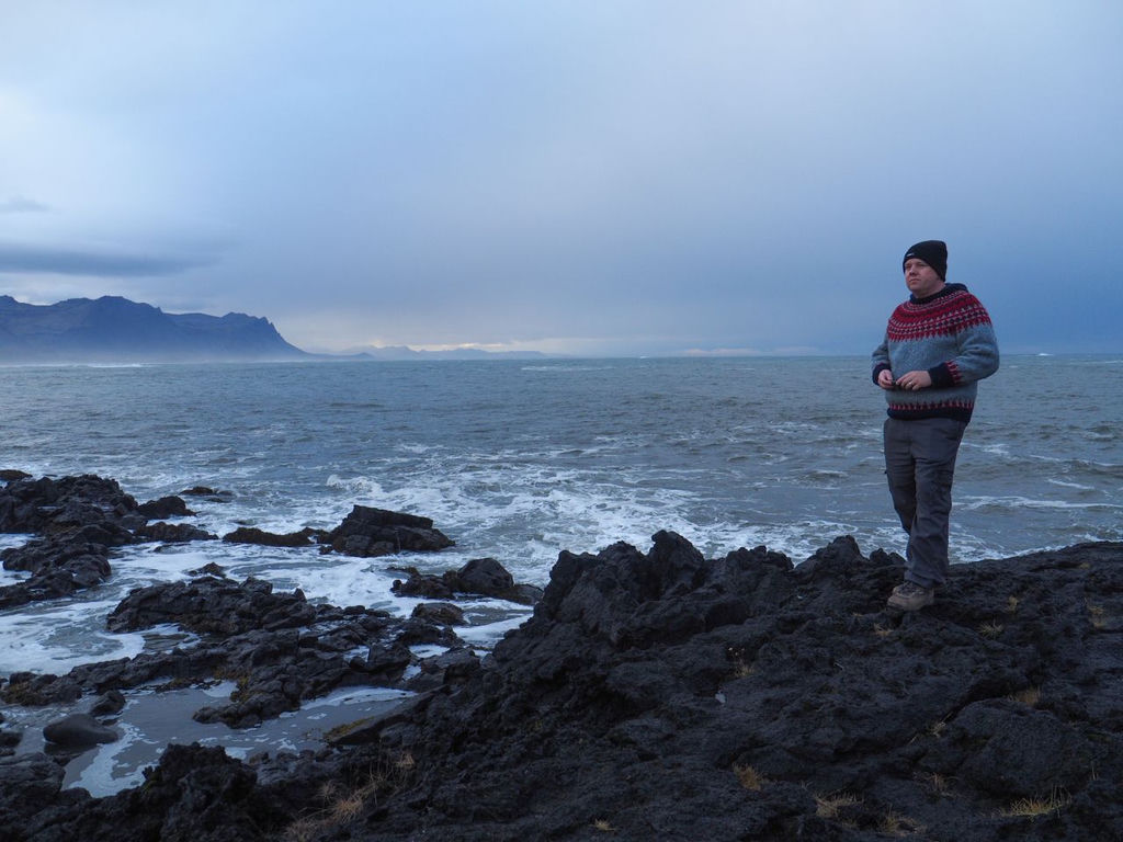 A man in an Icelandic jumper standing on a lava rock by the sea on a gloomy, stormy day.