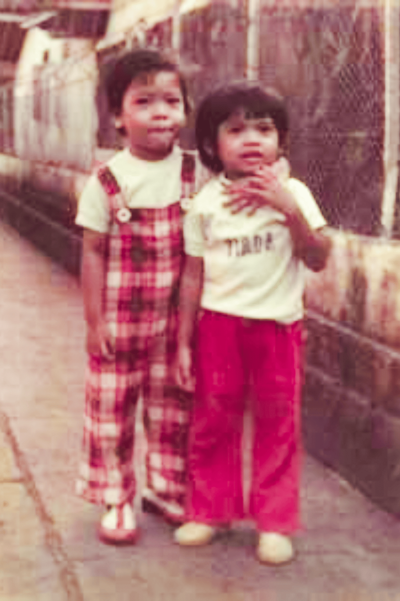 A little boy in a red, plaid dungaree and white t-shirt, and a little girl in a white t-shirt and red flare pants, his arm is around her shoulder, posing in an alleyway.